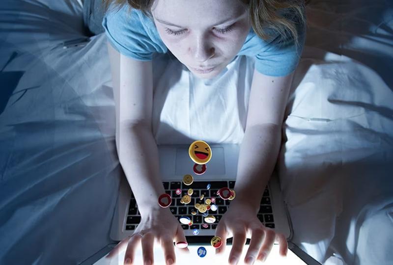 Young girl in bed on her laptop. Various emojis appear to be spilling out of the screen into her hands.