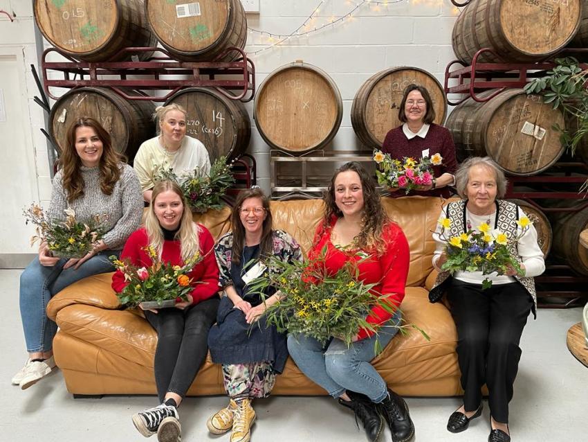 6 women sitting on sofa holding floral designs they have made, in front of barrels