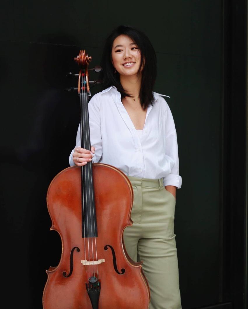 Cellist Rachel Siu in a white shirt and olive trousers, holding a cello and smiling