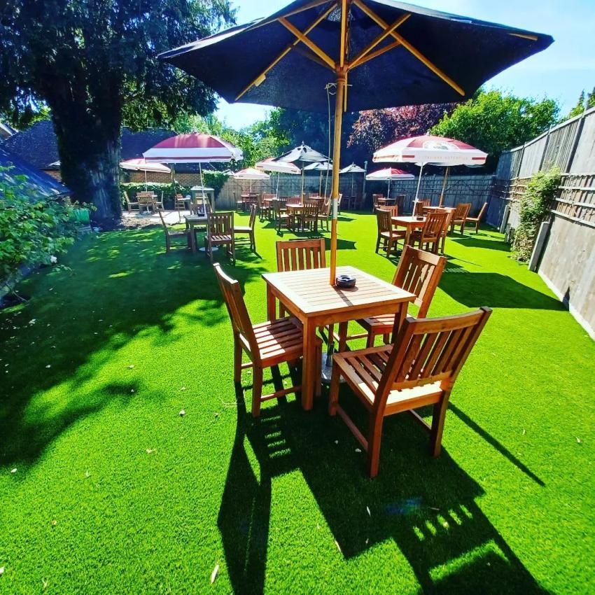 The beer garden at the wheelrights arms pub . green grass , lovely wooden furniture with shade from the parisols on a sunny day .