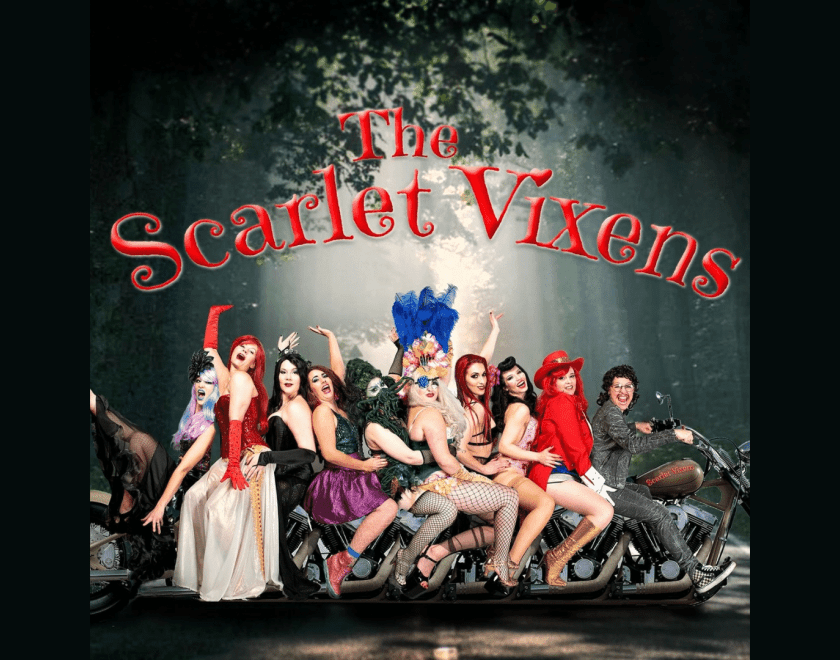 The Scarlet Vixens sit on a motorcycle smiling and laughing 