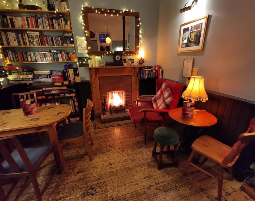 Corner of a room, with a lit fire in a hearth on the back wall. There is a mirror above the fire and a built-in bookcase beside it. In the middle ground are tables and chairs either side.s