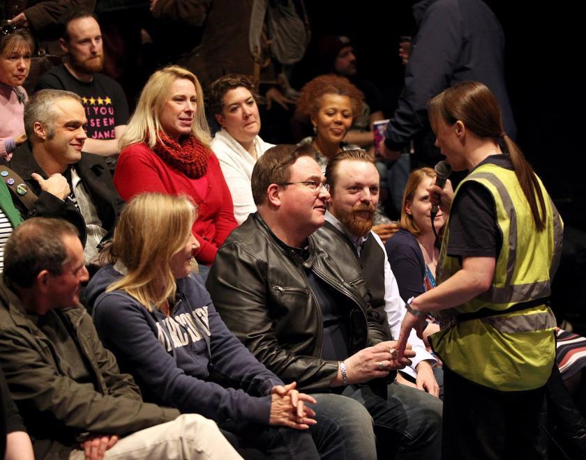 Photograph of an audience watching a performer wearing a hi-vis vest holding a microphone