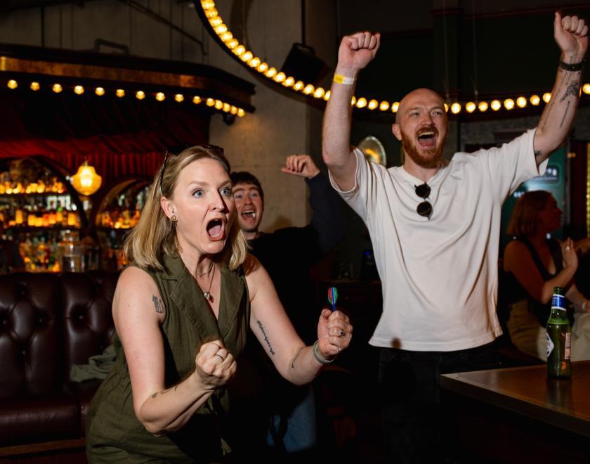 A woman and a man in a bar, gesturing their arms to denote success. She is holding a dart.