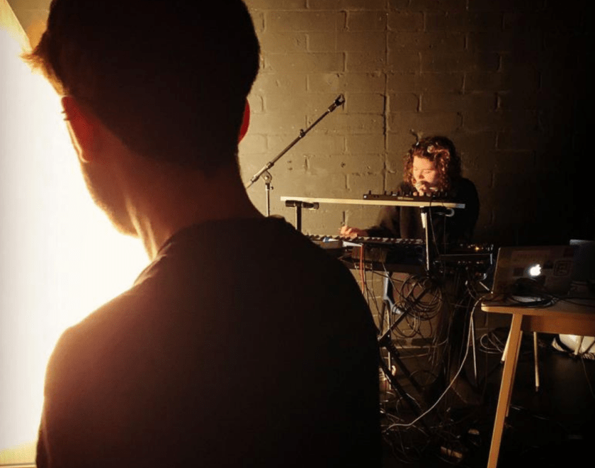The Dark rehearsal image 3 - in the foreground a performer stands in silhouette with their back to us, in the background we can see another performer sitting at an electronic keyboard