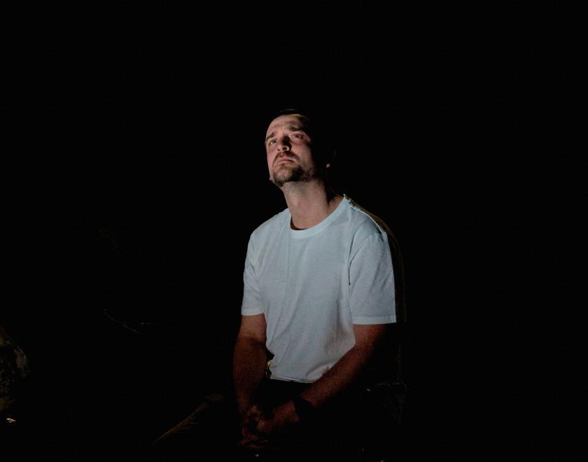 A performer in a plain white t-shirts sits on a stage in very low light, looking contemplative.