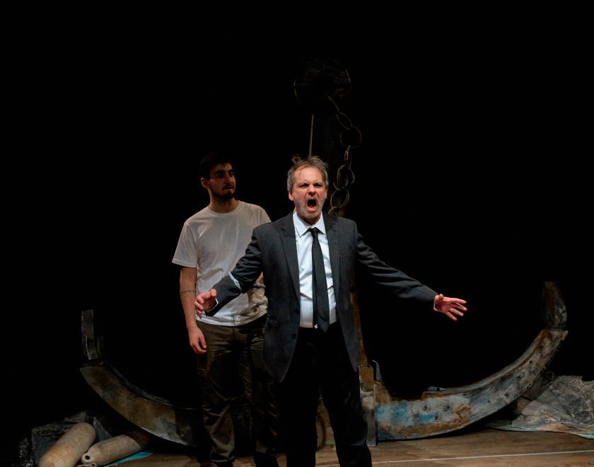 A performer in a suit and tie stands in the middle of the stage holding their arms open and looks as if they are screaming or shouting. Another performer in a white t-shirt stands close behind them.