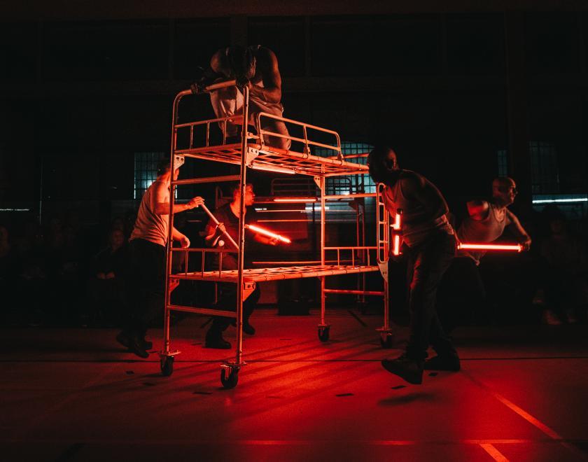 Photo of a prison bunk bed on wheels, with performers standing or crouching on it and around it. 
