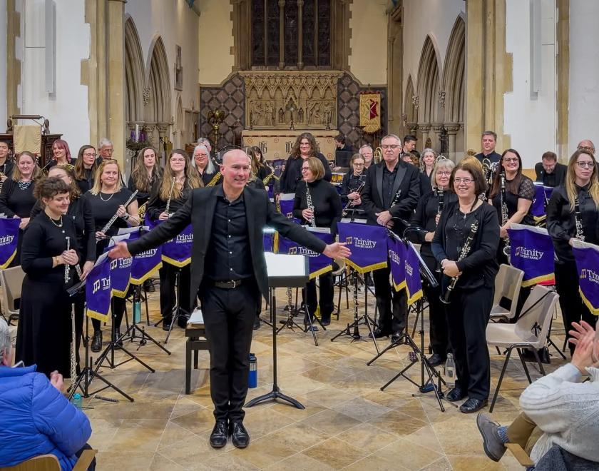 A conductor in black stands at the front of Trinity Concert Band inside a church, arms open as the audience applauds. Musicians dressed in black hold instruments behind purple Trinity-branded stands, with church arches and an ornate altar visible in the background.