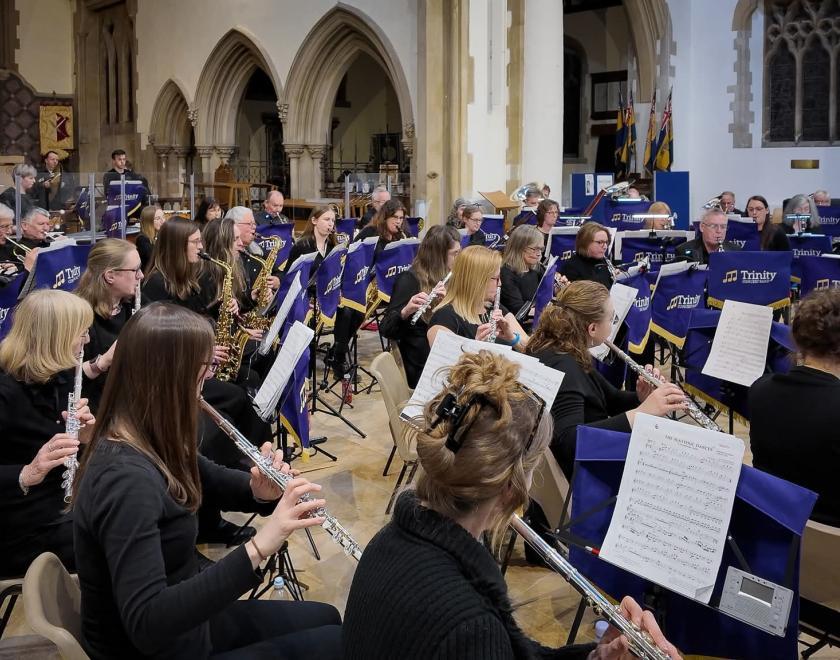 A side view of Trinity Concert Band performing inside a church, with flutes and saxophones in the foreground. Musicians dressed in black read from sheet music on purple Trinity-branded stands, while brass and percussion players sit further back beneath stone arches and church details.