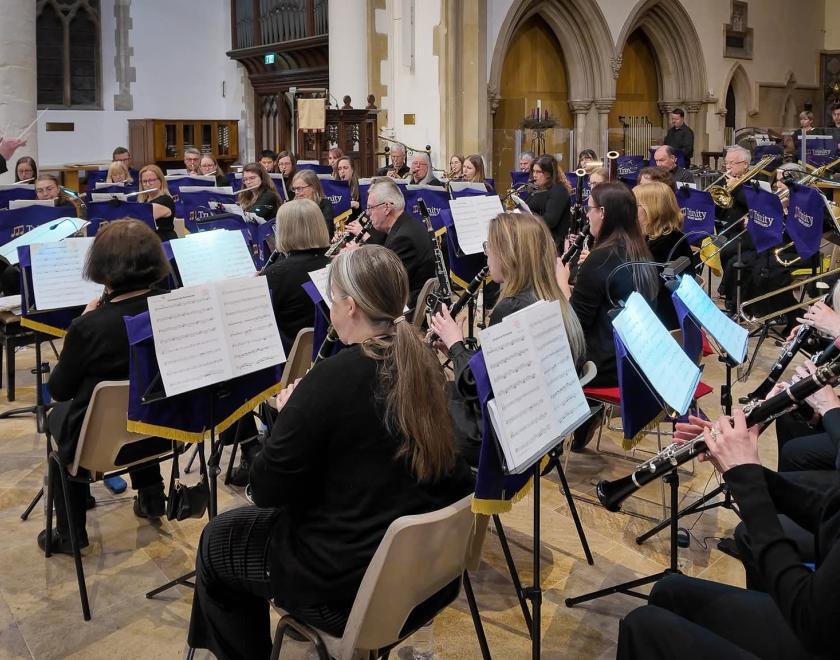 A wide side view of Trinity Concert Band performing inside a church, with the conductor at the front leading musicians in black. Clarinets and other woodwinds are in the foreground reading sheet music on purple Trinity-branded stands, while brass and percussion sections play behind under high stone arches and church interior features.