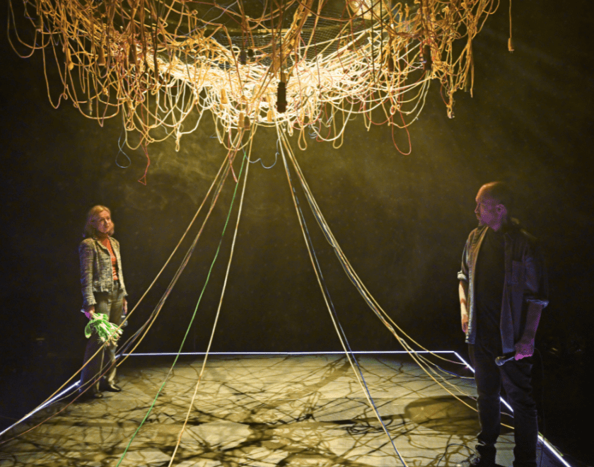 Production photo: A man and a woman stand on opposite sides of the stage. She is holding a bunch of flowers in one hand. The mass of wires suspended in the middle of the space has a number of wires hanging down and spread throughout the space.