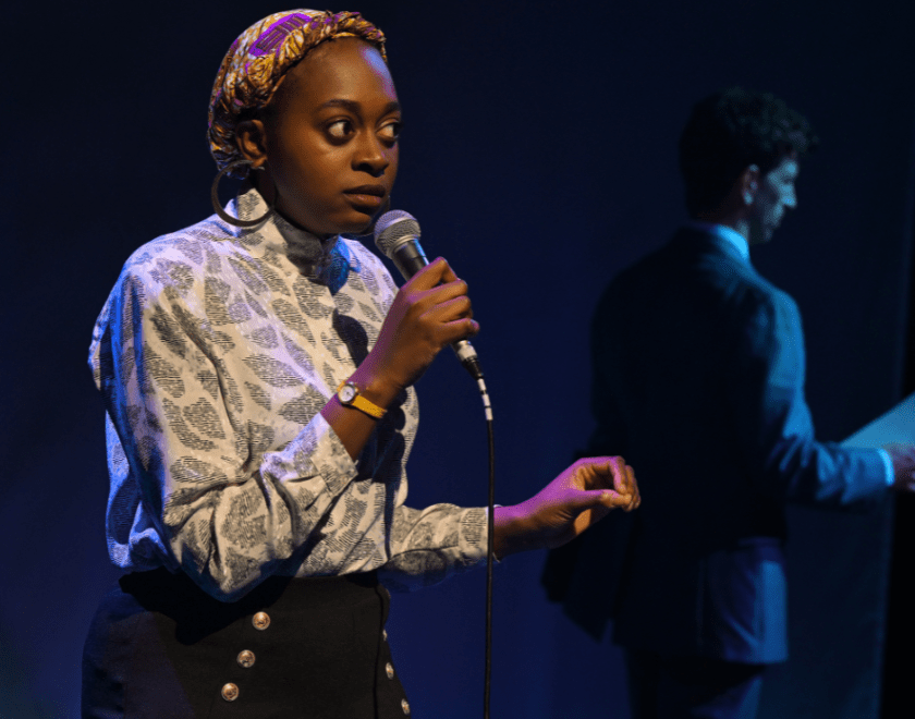 Production photo: A woman speaking into a microphone, a man is visible in the background