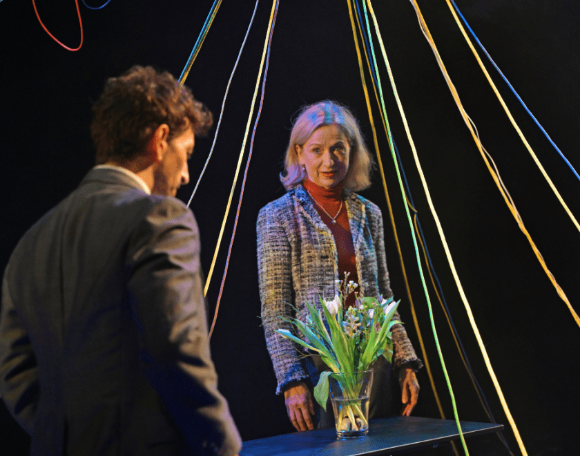 Production photo: A man and woman stand facing each other with multi-coloured wires hanging down around them and a vase of flowers in between them
