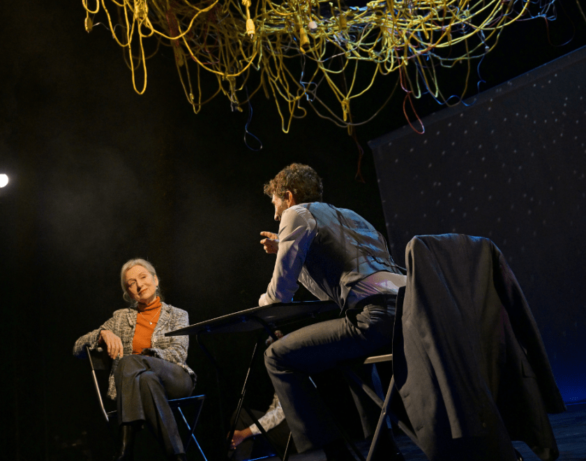 Production photo: Another angle of the man and woman at a table with wires suspended above them