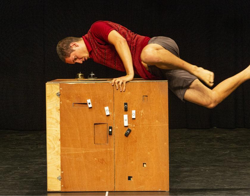 Production photo of Hermit. A performer wearing a red shirt and grey shorts appears to leapfrog over a wooden box with lots of doorbells attached to the front.