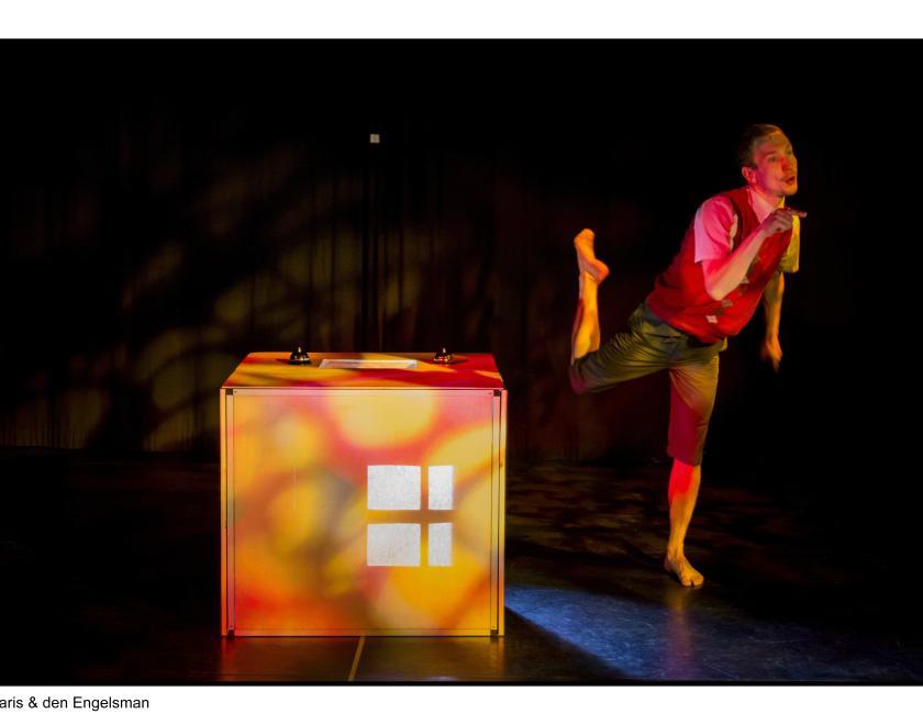 Another production photo. The same performer stands on one leg next to the big wooden box. The box has light projected on to it to make it look like it has a window.