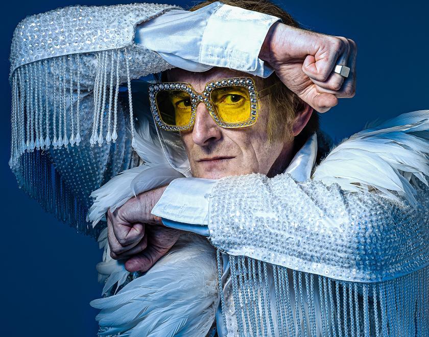 Photo of Henry Naylor dressed as Elton John, posing in front of a blue backdrop.