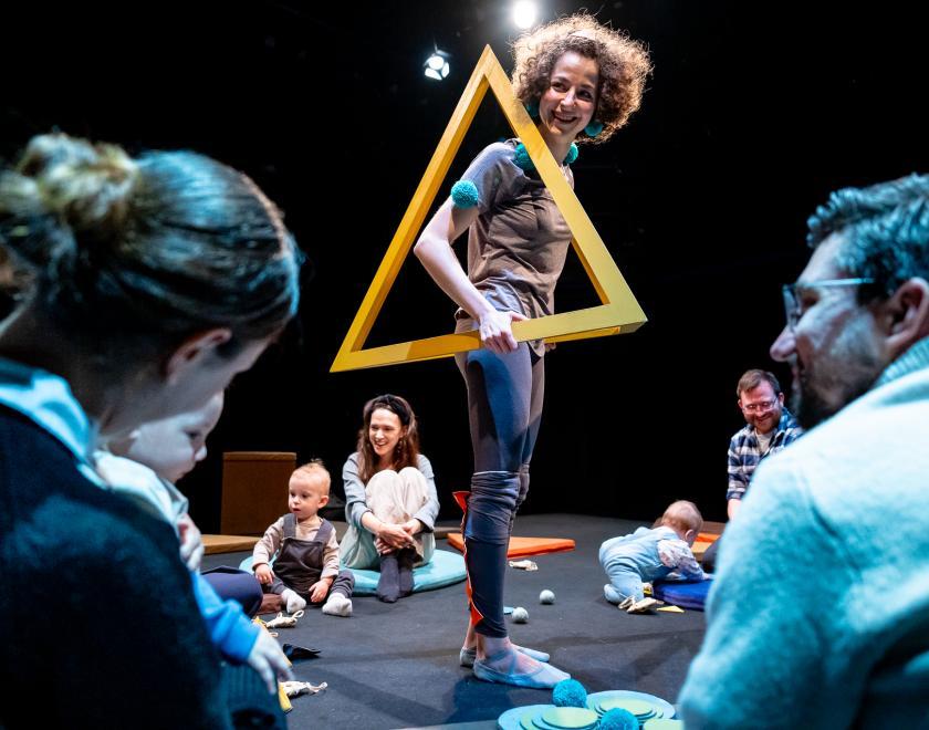 Production photo from 'Soft or Spiky?'. A performer stands in the middle of the performance space, holding a light orange pyramid-shaped frame around their torso with their head sticking out of the top. An audience of smiling adults and babies sits on the floor surrounding the performer.