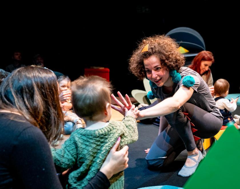Photo of the performer interacting with a baby in the audience, holding out their hand as the baby touches their fingers. Other adults and babies in the audience are visible in the background.