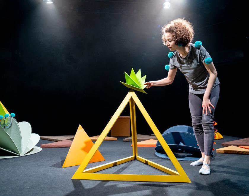 Photo of the performer standing next to a yellow pyramid shaped frame, and placing a smaller green spiky shaped prop on top of it.
