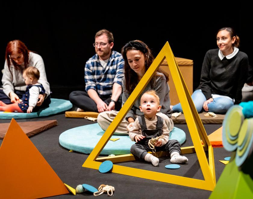 Photo of an audience of adults and babies, with various colourful prop shapes around them. One baby in the foreground is sitting inside the yellow pyramid-shaped frame and looks mesmerised by it.