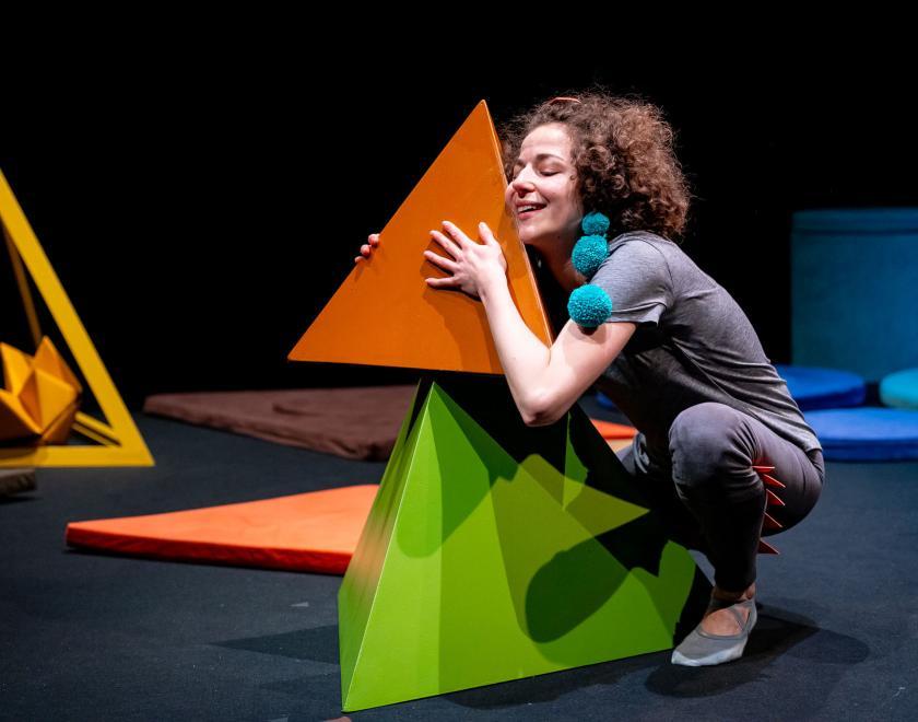 Photo of the performer squatting in the centre of the space with a big green pyramid shape in front of them, holding a smaller orange pyramid up to their face as if cuddling it. Other shapes in blue, orange and yellow are scatted around the space.