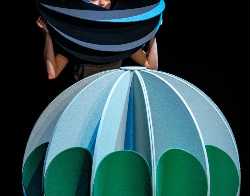 Photo of the performer standing behind a large green sphere prop with smaller green circles attached to it. The performer is holding a smaller blue sphere prop in front of their face and looking through a gap in it.