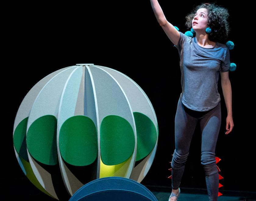 Photo of the performer standing next to a large green sphere prop, holding a small blue ball prop up to the light.
