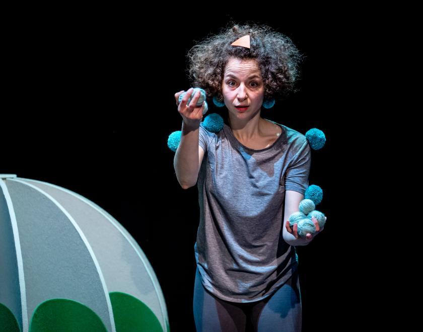 Photo of the performer standing next to a large green prop shape (not enough of it is visible to tell what it is) and holding a number of blue fluffy balls. They look like they are about to start juggling them.