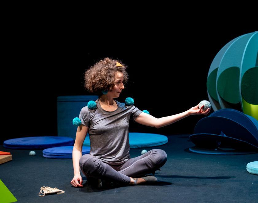 Photo of the performer sitting crossed-legged on the floor of the space, holding out a blue fluffy ball as if offering it to someone out of frame.