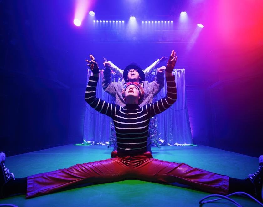 Photo of three cast members performing a scene, in a line one behind the other, on a stage lit with bright colours.