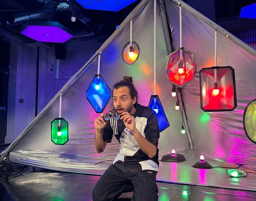 Photo of a performer holding a pair of multi-coloured spectacles in their hands in front of the set for 'Kaleidoscope'