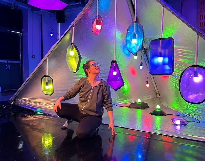 Photo of a performer wearing multi-coloured spectacles crouched in front of the set for 'Kaleidoscope' which is a large triangle tent with bright multi-coloured lights suspended from it.