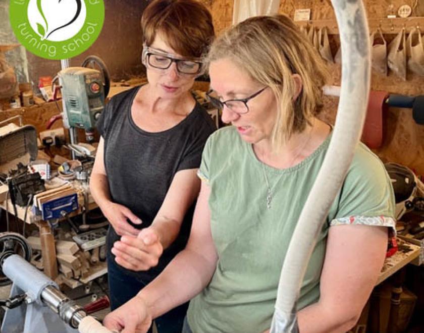 Liz Pearson teaching woodturning to a woman at the lathe.