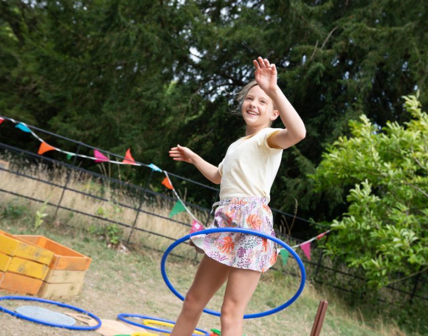 A young girl enjoying Summer of Play at Basildon Park. She is hoola hooping against a backdrop of parkland and bunting is hung along the play area.