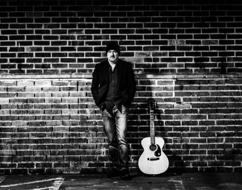 Photo of Craig Joiner with his guitar. Craig is standing in front of a wall in an urban street. The sky is dark and Craig is lit up by a street lamp. The photo is in black and white, giving it a retro feel.