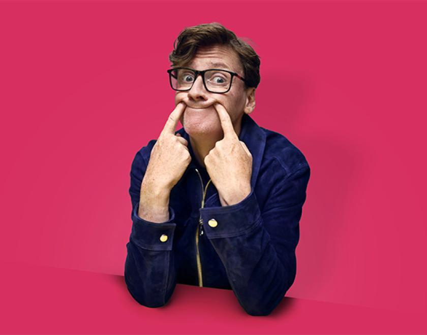 Photo of Ed Byrne overlaid onto a fuchsia pink background, pushing the corners of his mouth up with his fingers to force a smile