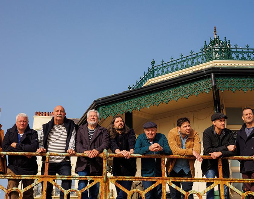 Photo of Fisherman's Friends standing in a row behind a railing, with the roof of a bandstand visible in the background.