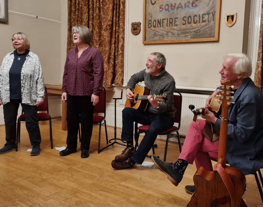 Photo of the four members of Four Gone Confusion, singing and playing musical instruments in a folk club. The band are dressed informally.