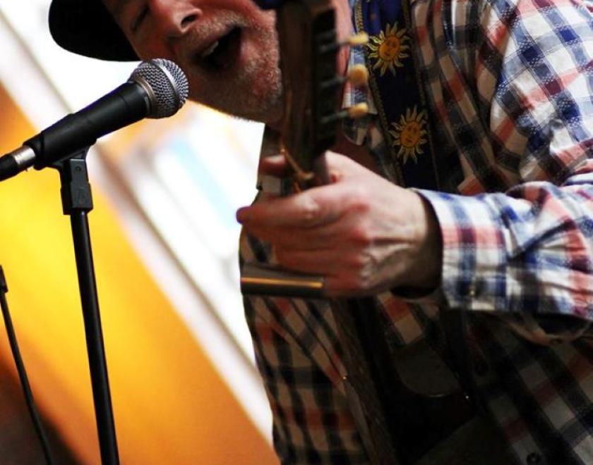 Older white man in a black hat and checked shirt plays a stringed instrument in front of a microphone