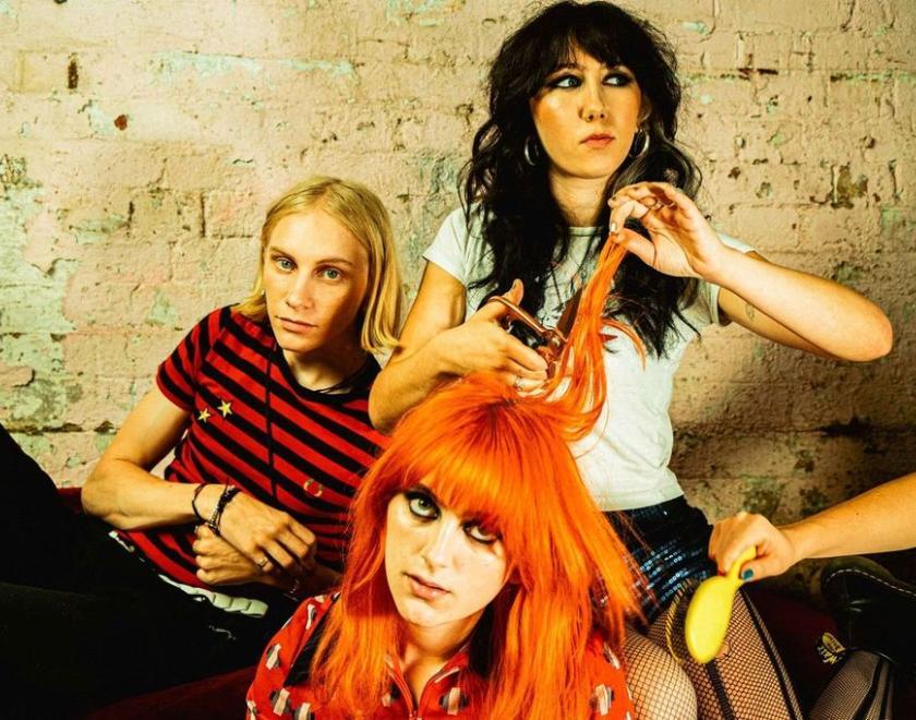 band pic of the trio. red head at front with brunette singer cutting their hair as the drummer looks on