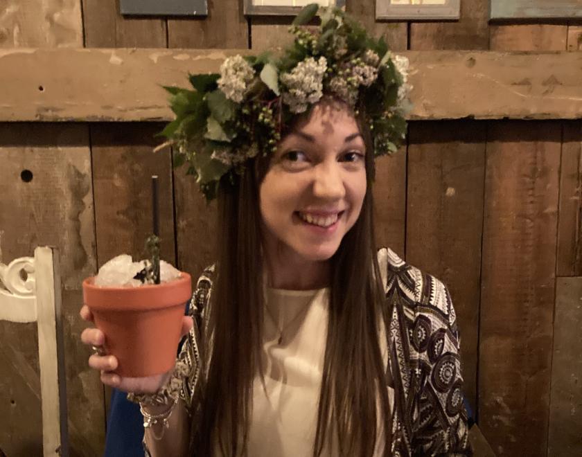 30-something woman wearing crown of foliage, frothy white flowers and  green berries raising a plantpot cocktail and smiling
