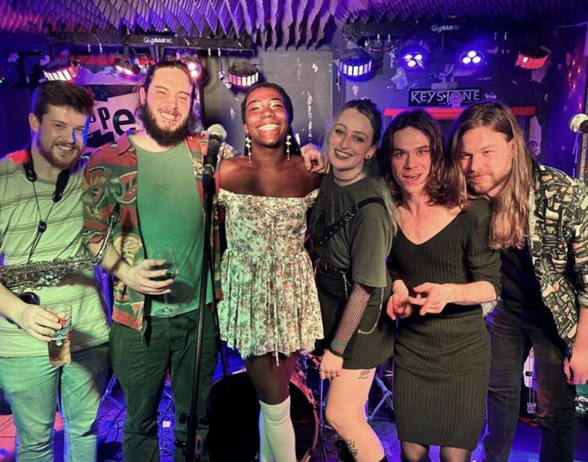 Six musicians with big smiles are standing leaning in towards each other facing the camera. They have blue lights overhead, three microphones in front and an amp to one side.