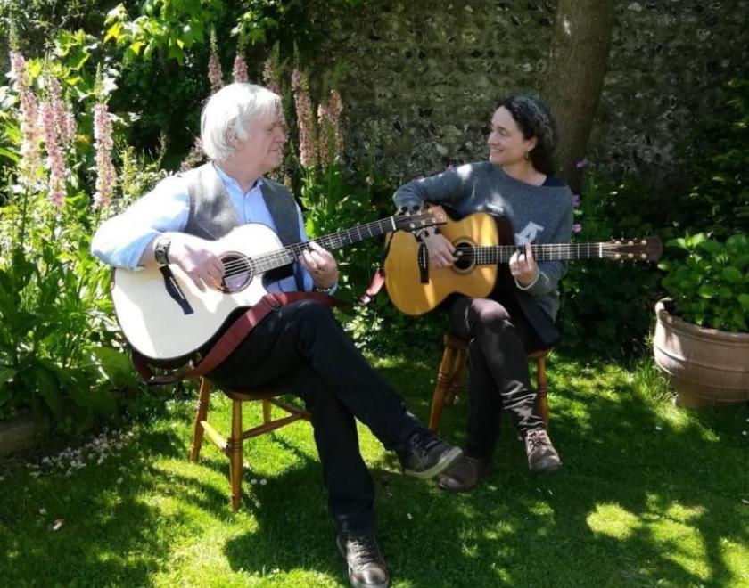 Photo of Natasha Norodien and Terry Lees sitting in a garden playing guitars and singing.