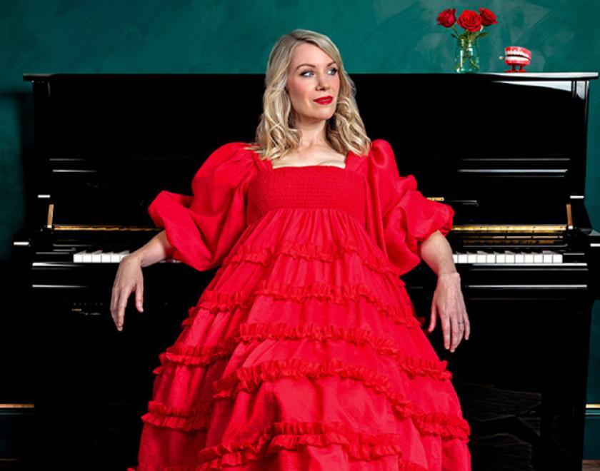 Photo of Rachel Parris wearing a red ballgown and sitting in front of a piano