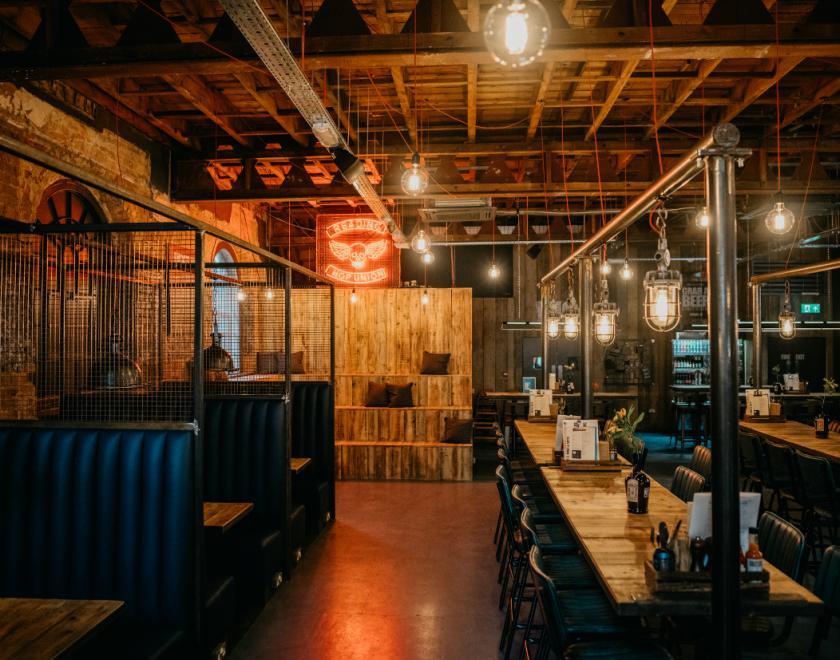 Bar seating area with a mix of high tables with stools and boothed seating. A red neon sign can be seen above wooden bleacher seating.