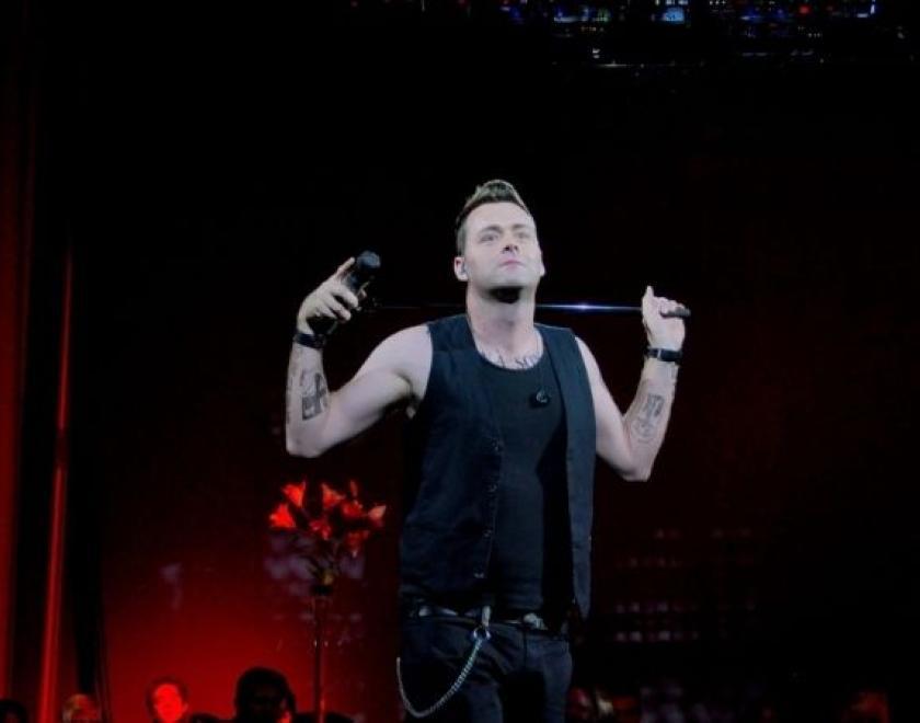 The image shows a Robbie William tribute artist stood in front of a red curtain.