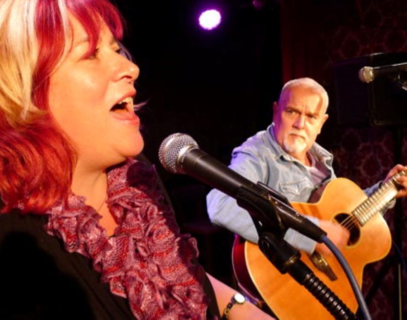 Sally Ironmonger and Brian Carter performing. Sally is singing and has a smile on her face. Brian is playing a guitar and looking towards Sally.