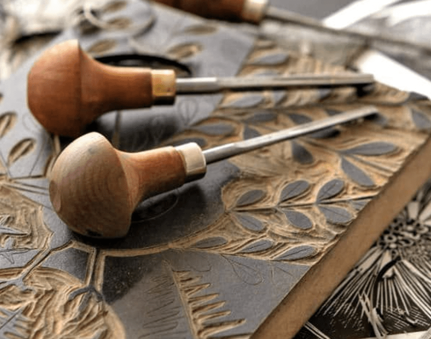 An image of a carved lino block with ink on. On top of the block are two carving tools with wooden handles.